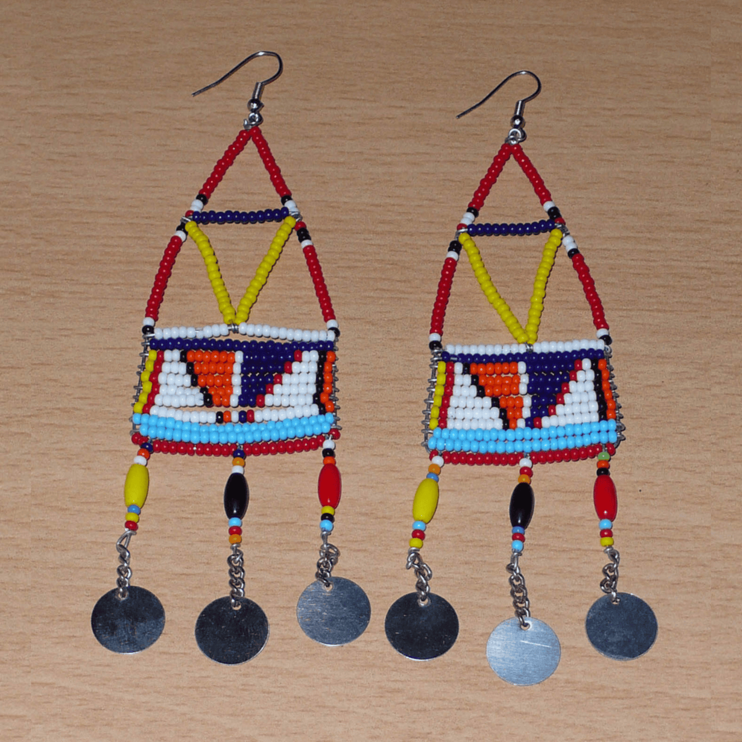 Longues boucles d'oreilles africaines à motifs géométriques Massai composées de perles de rocaille et de plastique rouges et multicolores, et de chaînettes décorées de perles et de disques métalliques. Elles mesurent 13 cm de long et 4 cm de large et se portent sur des oreilles percées.