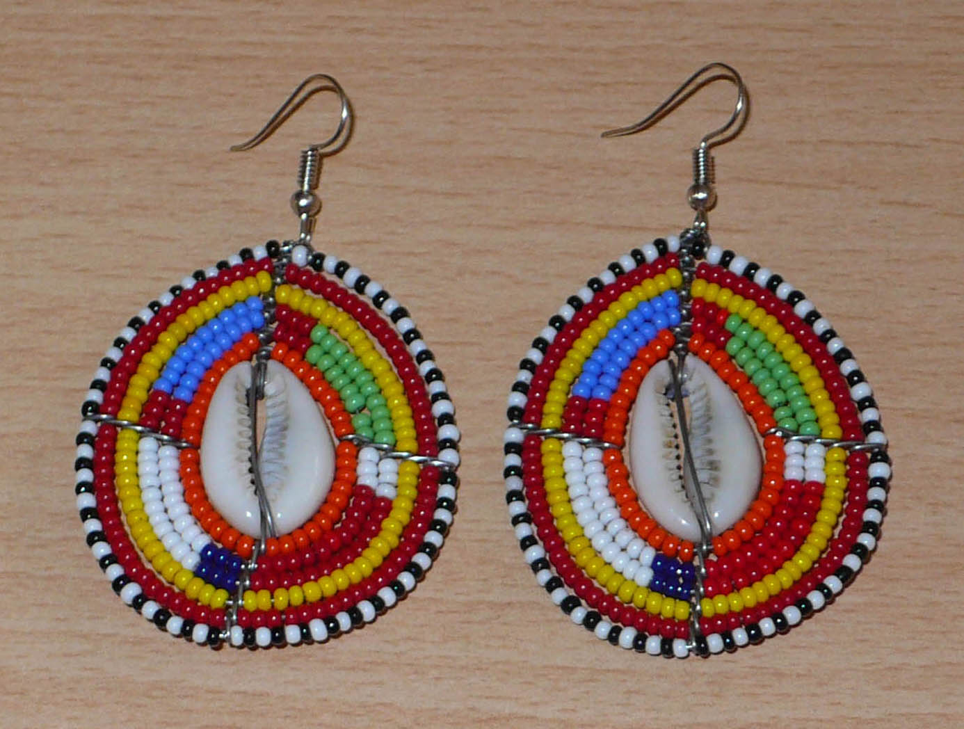 Boucles d'oreilles africaines ethniques composées de perles multicolores et de cauris montés en forme de boucliers Massai. Elles mesurent 7 cm de long et 4 cm de large et se portent sur des oreilles percées.