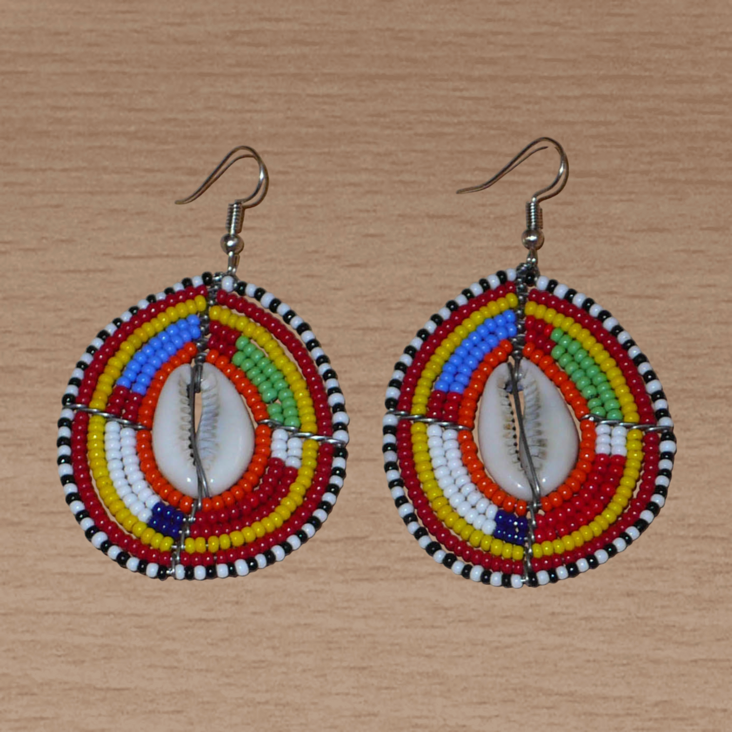 Boucles d'oreilles africaines ethniques composées de perles multicolores et de cauris montés en forme de boucliers Massai. Elles mesurent 7 cm de long et 4 cm de large et se portent sur des oreilles percées.