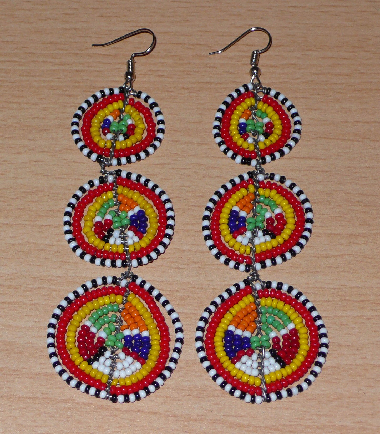 Affirmez votre style avec ces boucles d'oreilles africaines traditionnelles de confection artisanale Massai, évoquant le style afro chic des années 70. Elles se composent de perles de rocaille multicolores enfilées sur des fils de fer montés en forme de trois cercles superposés. Ces boucles d'oreilles mesurent 11 cm de long et 3.5 cm de large ; elles se portent avec des crochets en acier inoxydable sur des oreilles percées.