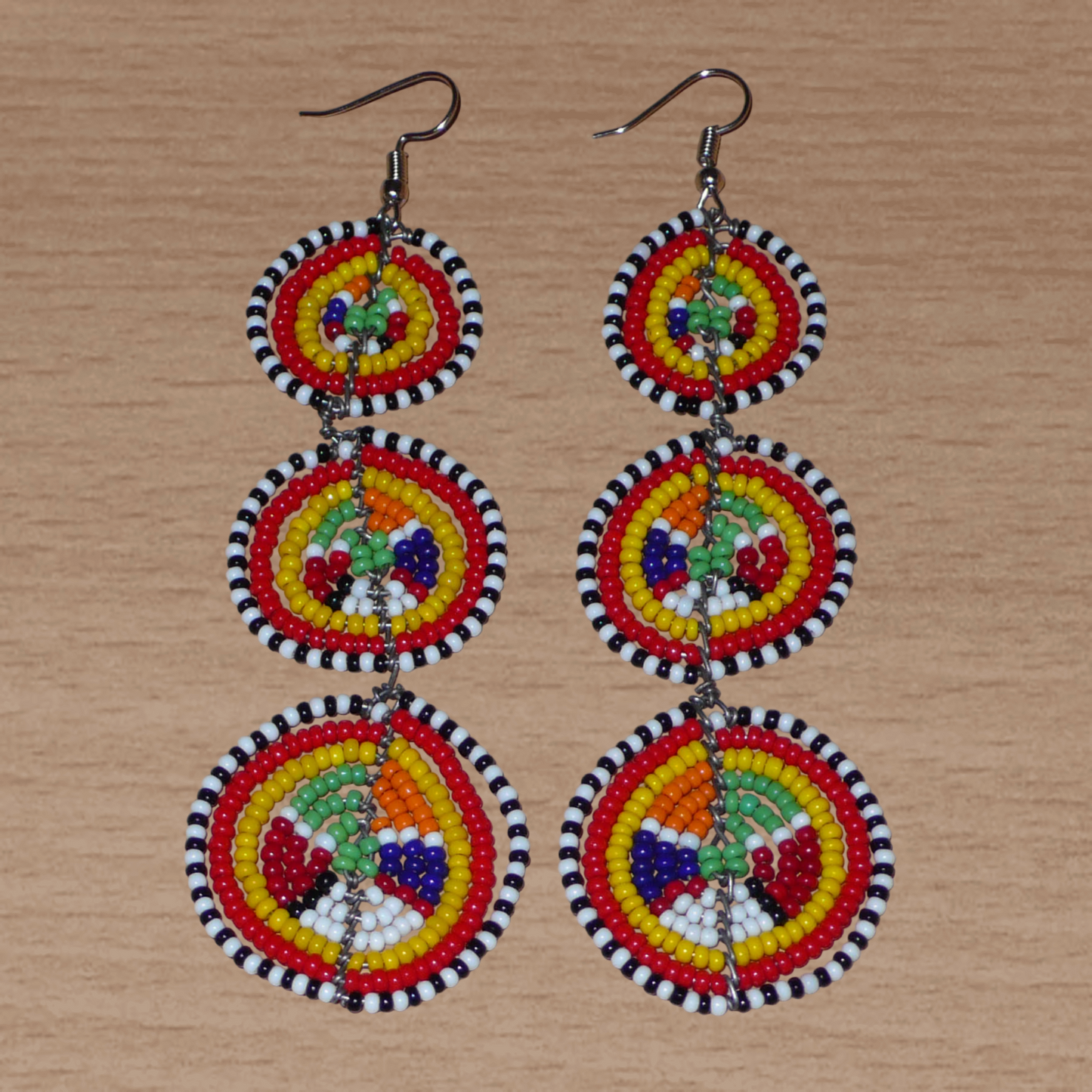 Affirmez votre style avec ces boucles d'oreilles africaines traditionnelles de confection artisanale Massai, évoquant le style afro chic des années 70. Elles se composent de perles de rocaille multicolores enfilées sur des fils de fer montés en forme de trois cercles superposés. Ces boucles d'oreilles mesurent 11 cm de long et 3.5 cm de large ; elles se portent avec des crochets en acier inoxydable sur des oreilles percées.