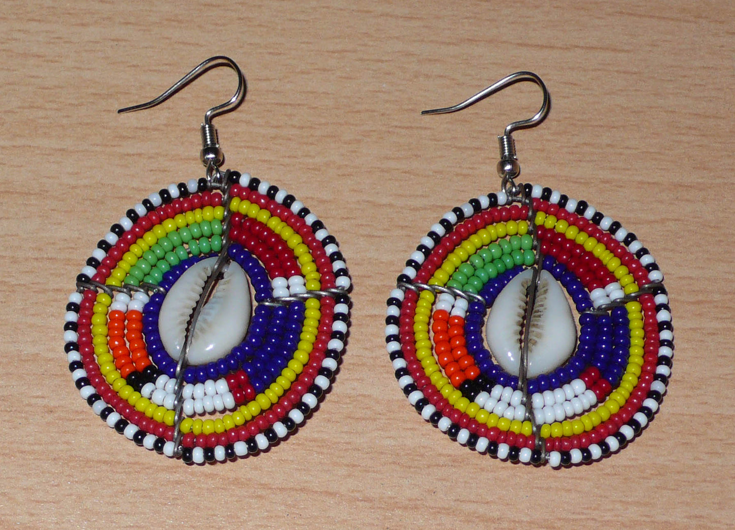 Boucles d'oreilles africaines traditionnelles aux motifs ethniques caractéristiques de l'artisanat Massai. De fabrication kenyane, elles se composent de perles de rocaille bleu foncé et multicolores et de cauris fixés sur des fils de fer montés en forme de boucliers Massai. Ces boucles d'oreilles de taille moyenne mesurent 6 cm de long et 4 cm de large ; elles se portent avec des crochets en acier inoxydable sur des oreilles percées.