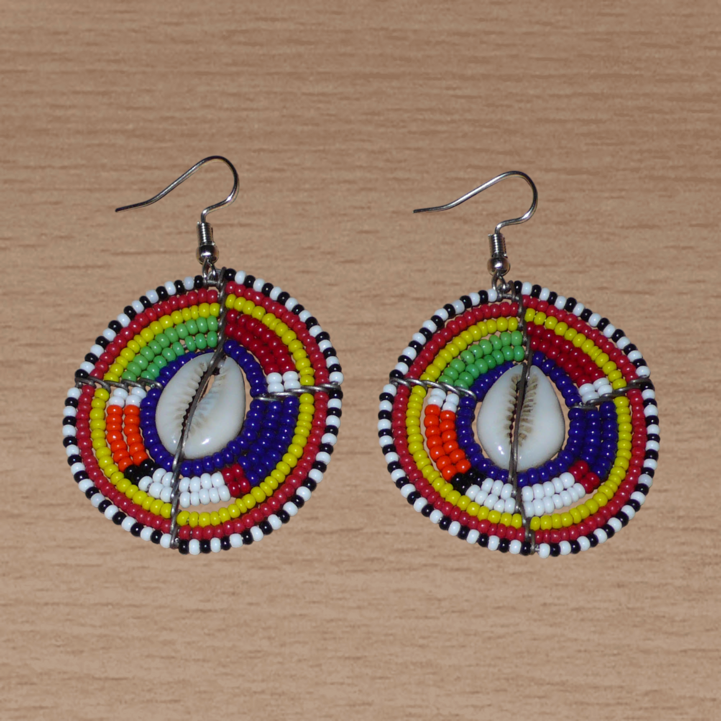Boucles d'oreilles africaines traditionnelles aux motifs ethniques caractéristiques de l'artisanat Massai. De fabrication kenyane, elles se composent de perles de rocaille bleu foncé et multicolores et de cauris fixés sur des fils de fer montés en forme de boucliers Massai. Ces boucles d'oreilles de taille moyenne mesurent 6 cm de long et 4 cm de large ; elles se portent avec des crochets en acier inoxydable sur des oreilles percées.