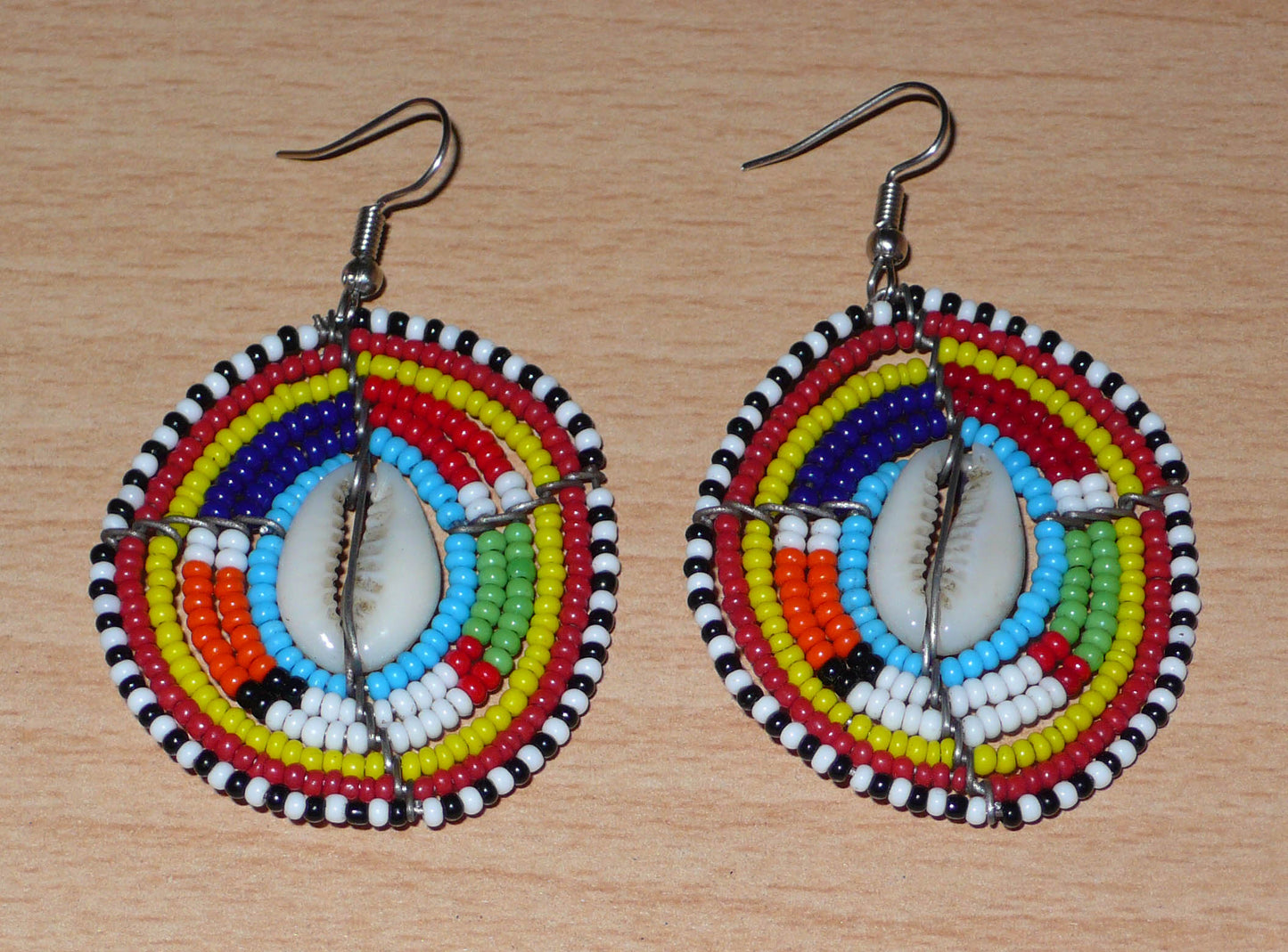 Boucles d'oreilles africaines traditionnelles aux motifs ethniques caractéristiques de l'artisanat Massai. De fabrication kenyane, elles se composent de perles de rocaille bleues et multicolores et de coquillages fixés sur des fils de fer montés en forme de boucliers Massai. Ces boucles d'oreilles de taille moyenne mesurent 6.5 cm de long et 4 cm de large ; elles se portent avec des crochets en acier inoxydable sur des oreilles percées.