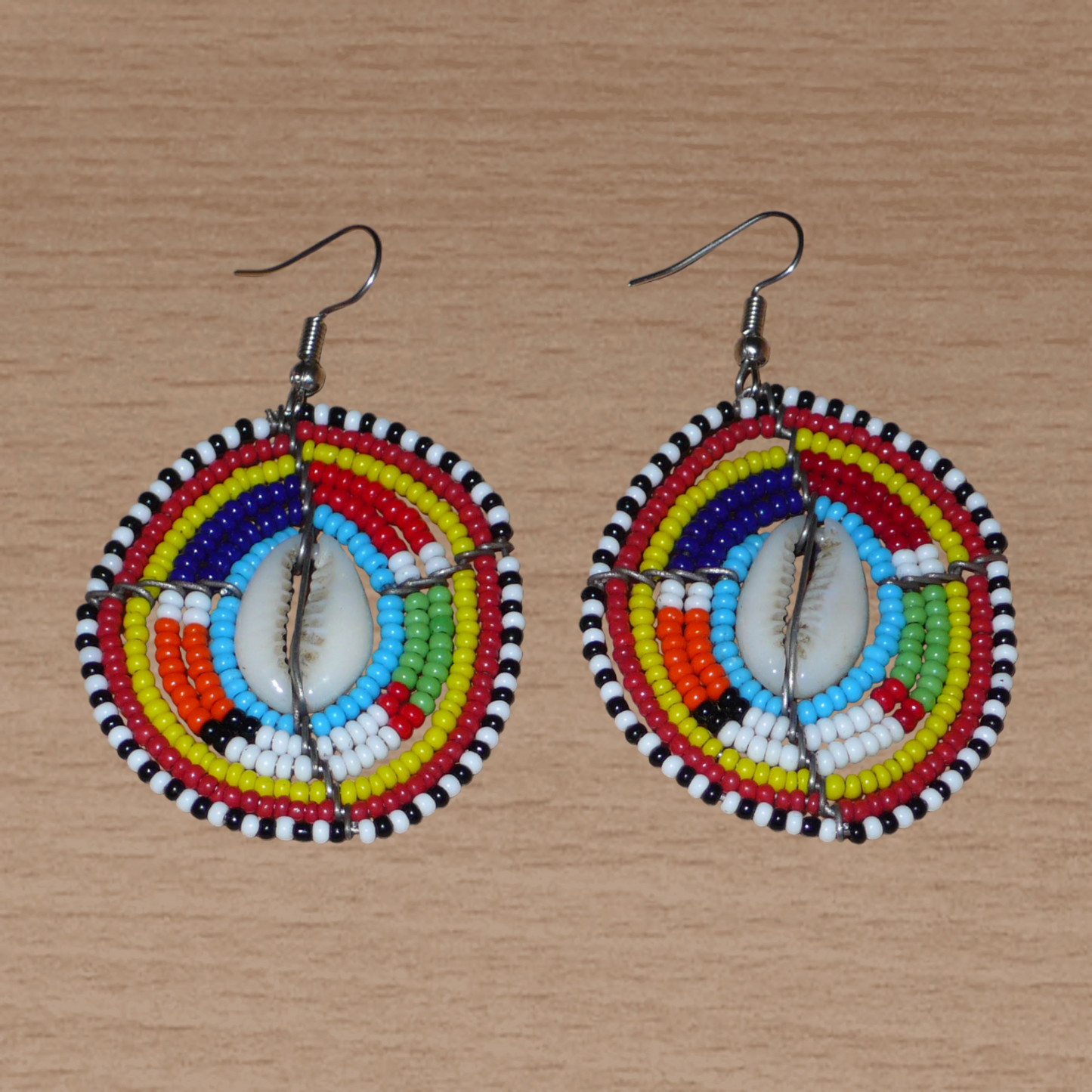 Boucles d'oreilles africaines traditionnelles aux motifs ethniques caractéristiques de l'artisanat Massai. De fabrication kenyane, elles se composent de perles de rocaille bleues et multicolores et de coquillages fixés sur des fils de fer montés en forme de boucliers Massai. Ces boucles d'oreilles de taille moyenne mesurent 6.5 cm de long et 4 cm de large ; elles se portent avec des crochets en acier inoxydable sur des oreilles percées.