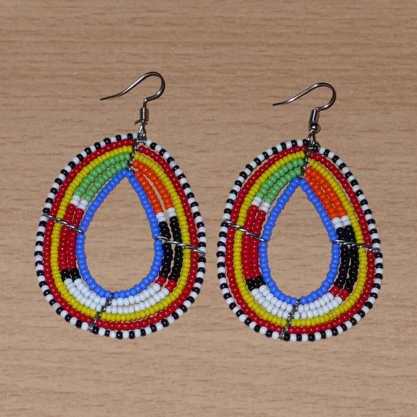 Boucles d'oreilles ethniques multi-rangs à motifs traditionnels Massai composées de perles de rocaille bleues et multicolores glissées sur six rangs de fils de fer montés en forme de gouttes. Elles mesurent 7.5 cm de long et 4 cm de large, et se portent avec des crochets en acier inoxydable sur des oreilles percées.