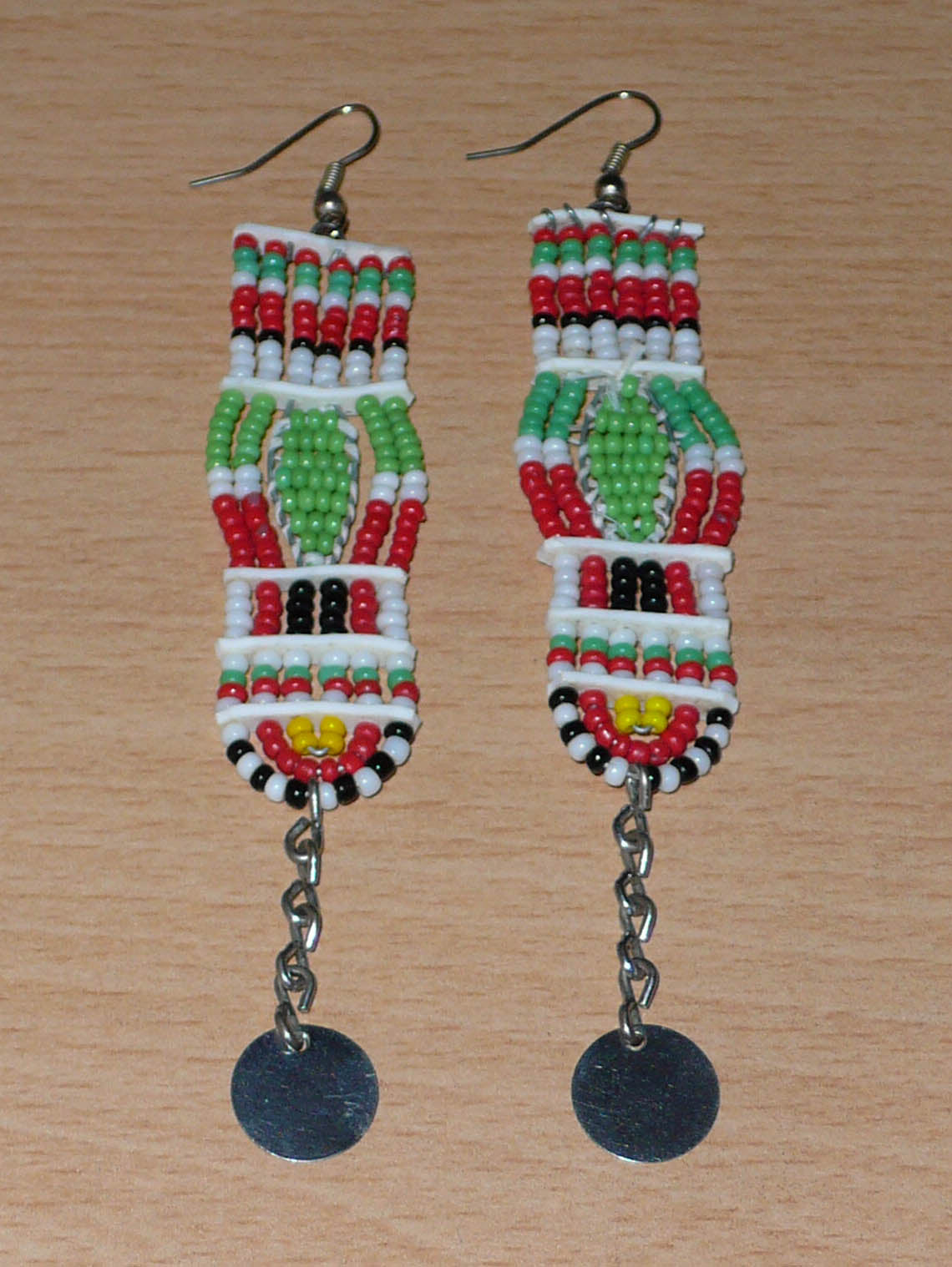 Longues boucles d'oreilles africaines de tradition Massai en perles de rocaille prolongées de chaînettes ornées de disques métalliques. Elles mesurent 11 cm de long et 2,5 cm de large, et se portent avec des crochets en acier inoxydable sur des oreilles percées.