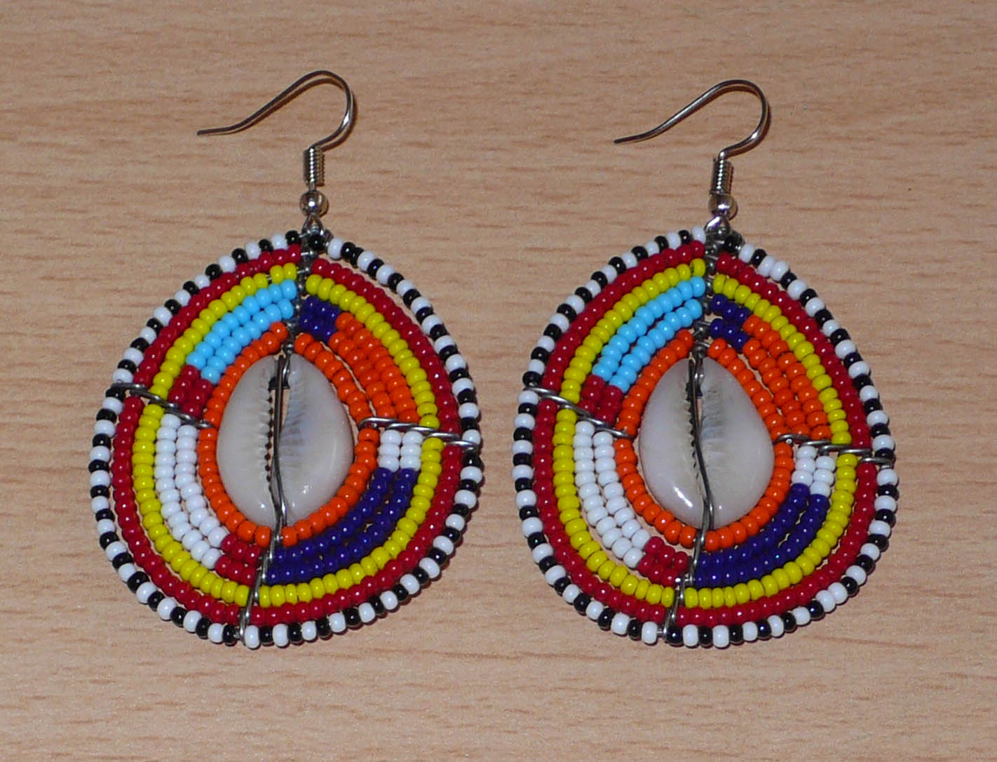 Boucles d'oreilles africaines Massai composées de perles de rocaille multicolores et de coquillages cauris. Elles mesurent 7 cm de long et 4 cm de large et se portent avec des crochets en acier inoxydable sur des oreilles percées.