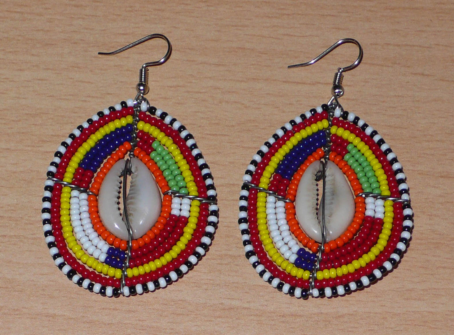 Boucles d'oreilles africaines à motifs ethniques composées de perles et de coquillages montés en forme de boucliers Massai. Elles mesurent 7 cm de long et 4 cm de large et se portent sur des oreilles percées.