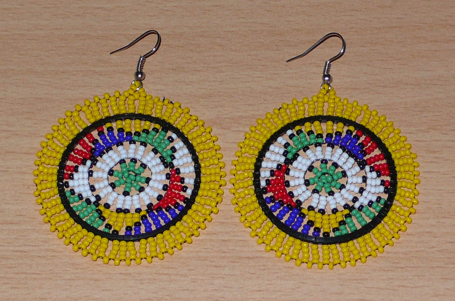 Boucles d'oreilles africaines zouloues en perles de rocaille jaunes et multicolores enfilées sur des structures circulaires en fils de fer et de nylon. Elles mesurent 7,5 cm de long et 5,5 cm de large et se portent avec des crochets en acier inoxydable.