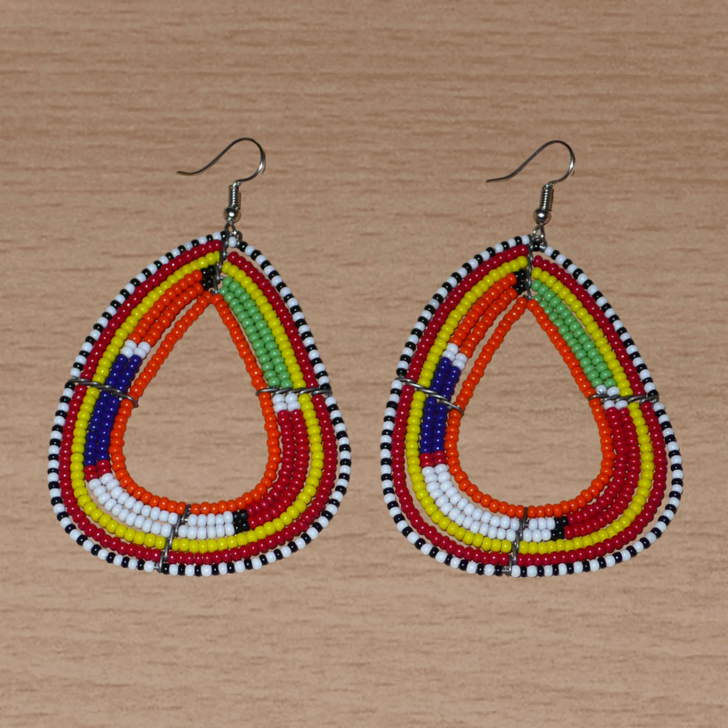 Boucles d'oreilles africaines traditionnelles à motifs ethniques Massai composées de perles de rocaille orange et multicolores enfilées sur six rangées de fils de fer montés en forme de triangles aux bords arrondis. Elles mesurent 8,5 cm de long et 5 cm de large et se portent avec des crochets en acier inoxydable sur des oreilles percées.
