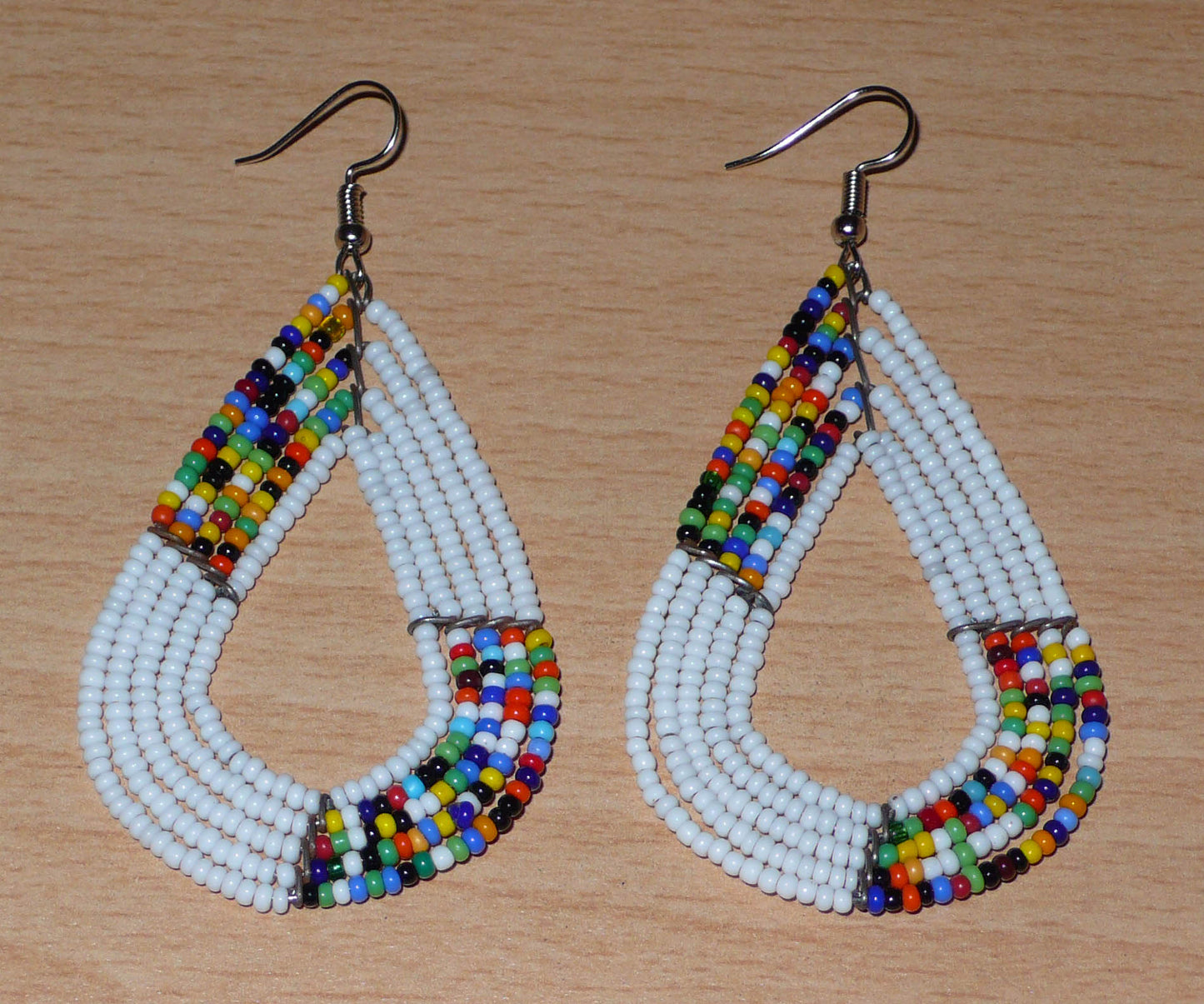 Boucles d'oreilles africaines multi-rangs blanches et multicolores aux accents évoquant le style bohème des années 70. Fabriquées au Kenya, elles se composent de perles de rocaille blanches et d'un mélange de perles multicolores glissées sur cinq rangées de fils de fer en forme de gouttes légèrement triangulaires. Ces boucles d'oreilles mesurent 8 cm de long et 4 cm de large ; elles se portent avec des crochets en acier inoxydable sur des oreilles percées.