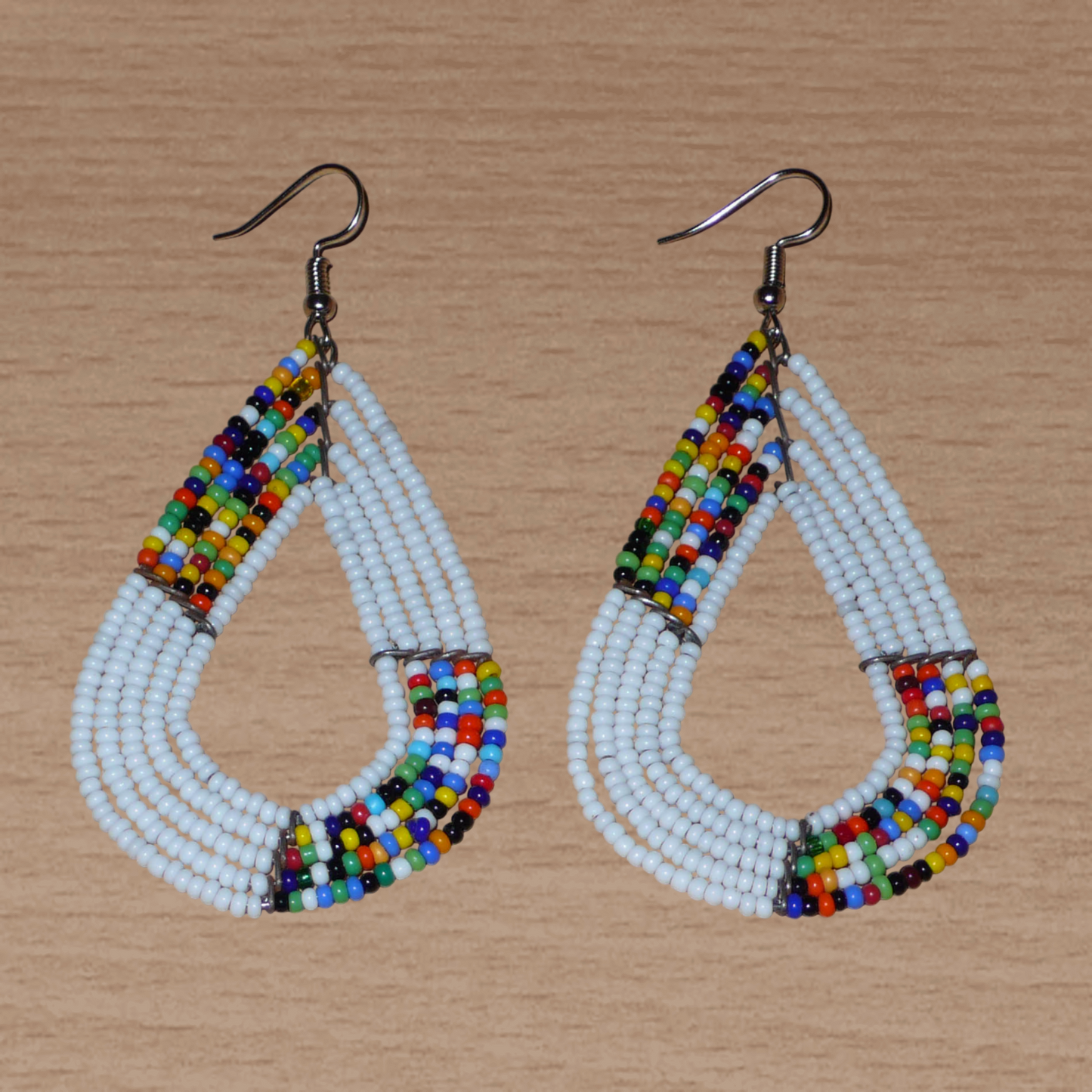 Boucles d'oreilles africaines multi-rangs blanches et multicolores aux accents évoquant le style bohème des années 70. Fabriquées au Kenya, elles se composent de perles de rocaille blanches et d'un mélange de perles multicolores glissées sur cinq rangées de fils de fer en forme de gouttes légèrement triangulaires. Ces boucles d'oreilles mesurent 8 cm de long et 4 cm de large ; elles se portent avec des crochets en acier inoxydable sur des oreilles percées.