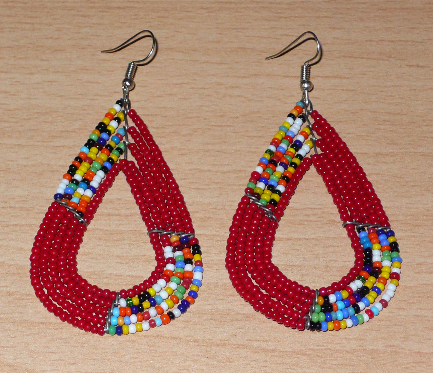 Boucles d'oreilles africaines Massai rouges et multicolores aux accents évoquant le style boho chic des années 70. Confectionnées au Kenya, elles se composent de perles de rocaille rouges et d'un mélange de perles multicolores glissées sur cinq rangées de fils de fer en forme de gouttes légèrement triangulaires. Ces boucles d'oreilles mesurent 8 cm de long et 4 cm de large ; elles se portent avec des crochets en acier inoxydable sur des oreilles percées.