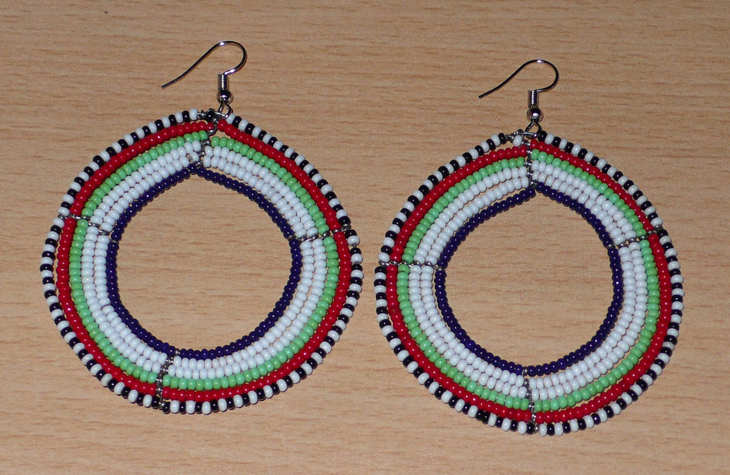 Boucles d'oreilles africaines Massai composées de perles de rocaille blanches, vertes, rouges, bleu foncé et noires glissées sur des fils de fer montés en forme de cercles. Elles mesurent 7 cm de long et 5 cm de large, et se portent avec des crochets en acier inoxydable sur des oreilles percées.