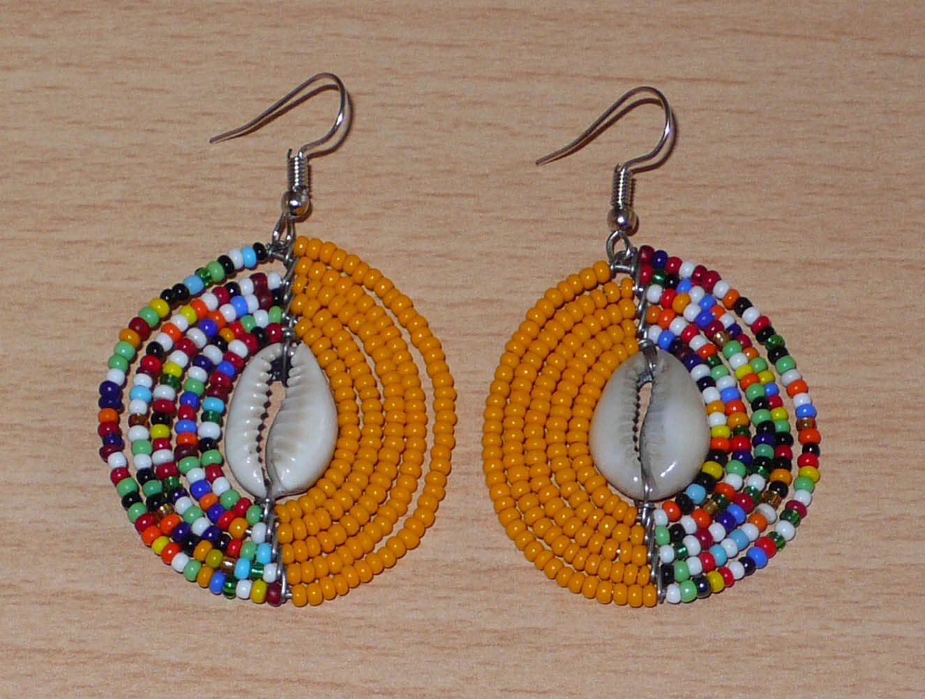 Boucles d'oreilles ethniques orange et multicolores fabriquées au Kenya composées de perles de rocaille et de coquillages cauris. De taille moyenne, elles mesurent 6 cm de long et 3,5 cm de large et se portent sur des oreilles percées.