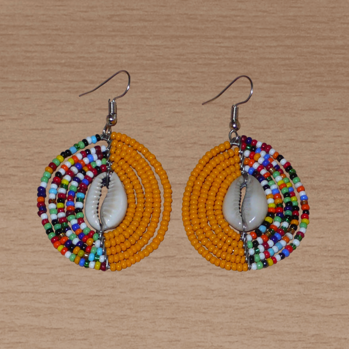 Boucles d'oreilles ethniques orange et multicolores fabriquées au Kenya composées de perles de rocaille et de coquillages cauris. De taille moyenne, elles mesurent 6 cm de long et 3,5 cm de large et se portent sur des oreilles percées.