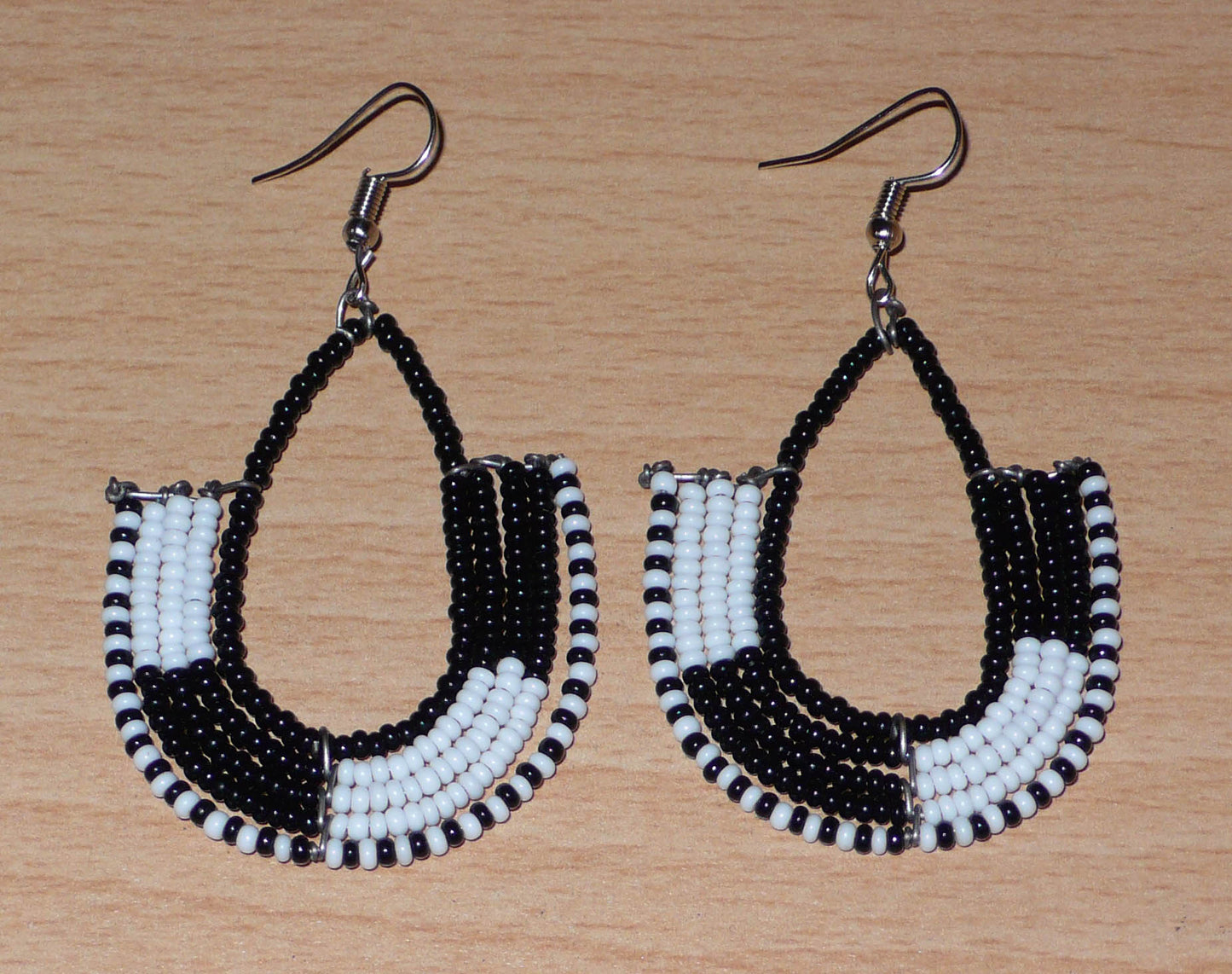 Boucles d'oreilles ethniques bicolores en forme d'ancres composées de perles de rocaille noires et blanches glissées sur des structures géométriques en fils de fer. De taille moyenne, ces boucles d'oreilles aux accents afro chic mesurent 7 cm de long et 4 cm de large ; elles se portent avec des crochets en acier inoxydable sur des oreilles percées.