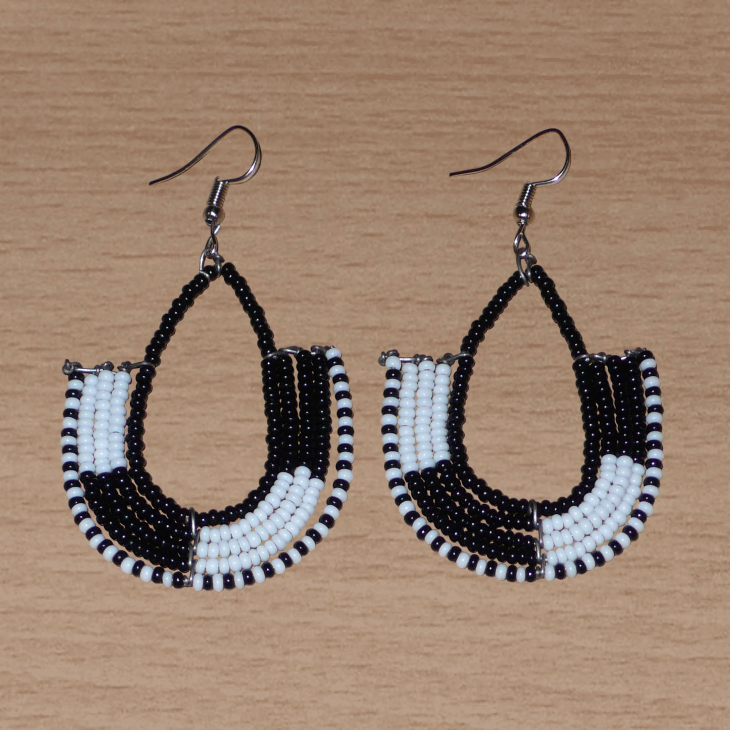 Boucles d'oreilles ethniques bicolores en forme d'ancres composées de perles de rocaille noires et blanches glissées sur des structures géométriques en fils de fer. De taille moyenne, ces boucles d'oreilles aux accents afro chic mesurent 7 cm de long et 4 cm de large ; elles se portent avec des crochets en acier inoxydable sur des oreilles percées.