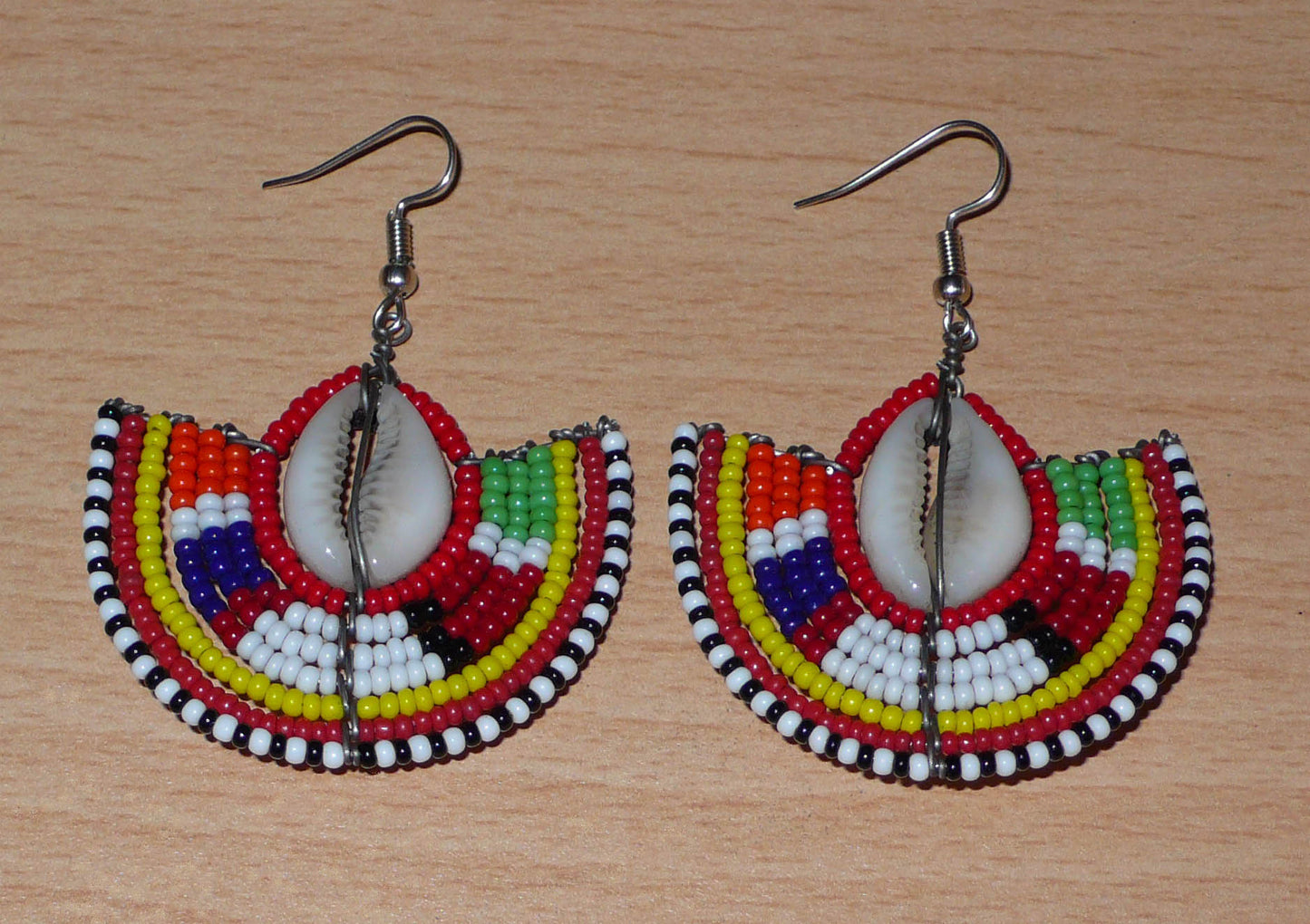 Ajoutez une touche exotique à votre style avec cette jolie paire de boucles d'oreilles africaines issues de l'artisanat traditionnel Massai. Ces boucles d'oreilles ethniques chic se composent de perles de rocaille rouges et multicolores et de coquillages cauris. De taille moyenne, elles mesurent 6 cm de long et 4.5 cm de large, et se portent avec des crochets en acier inoxydable sur des oreilles percées.
