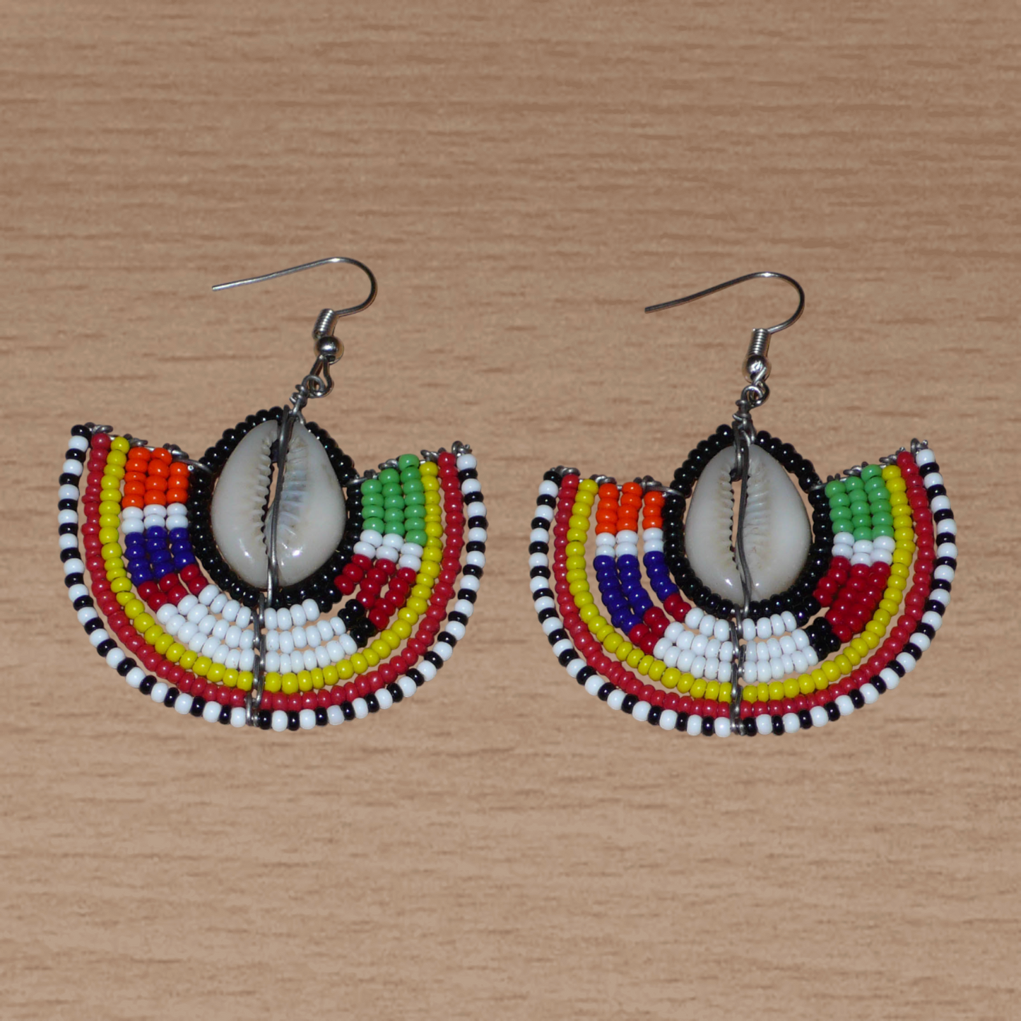 Apportez une note de couleurs à votre look avec cette paire de boucles d'oreilles africaines au style exotique chic issues de l'artisanat traditionnel Massai. Confectionnées à la main au Kenya, ces boucles d'oreilles se composent de perles de rocaille noires et multicolores et de coquillages cauris. De taille moyenne, elles mesurent 6 cm de long et 4.5 cm de large, et se portent avec des crochets en acier inoxydable sur des oreilles percées.