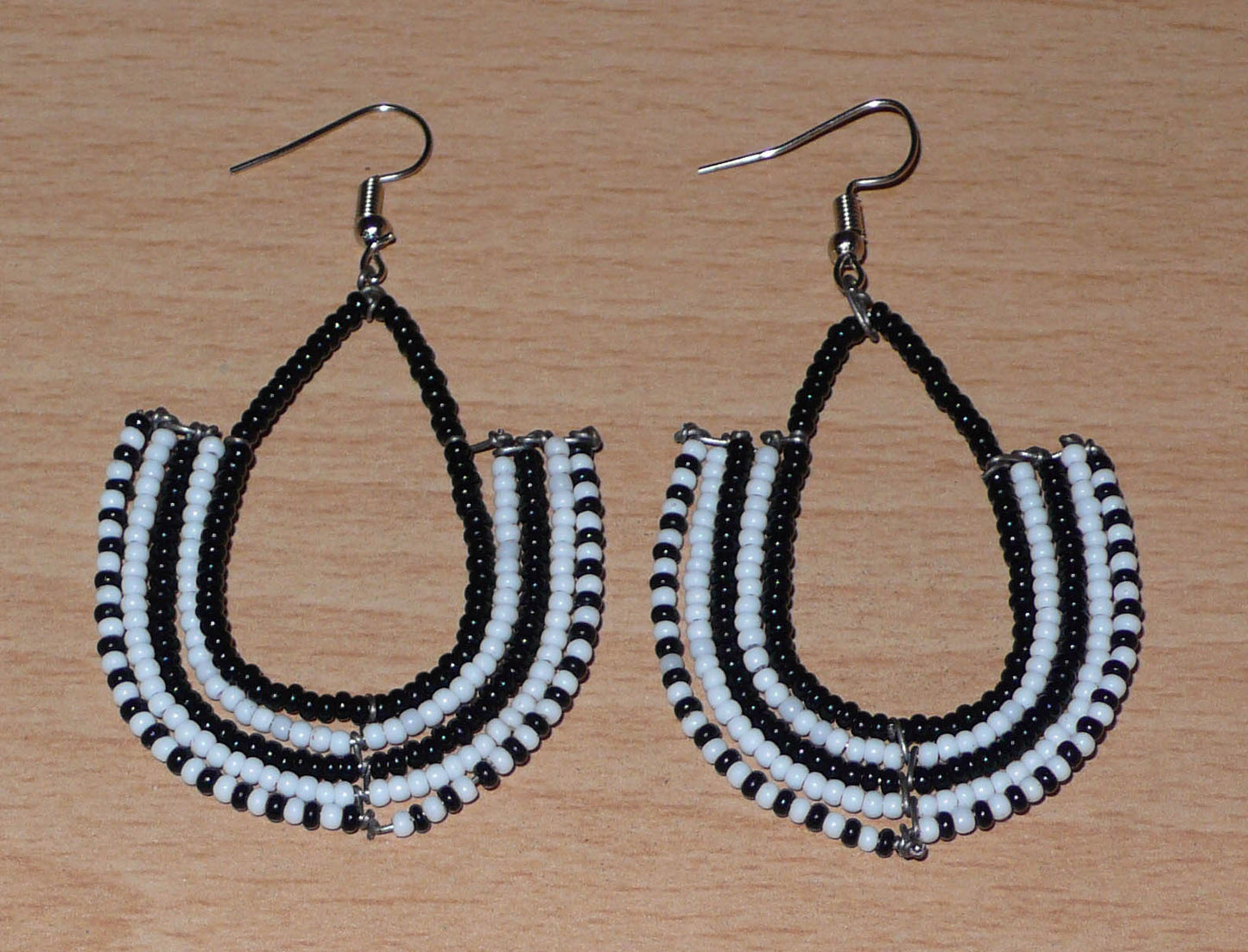 Boucles d'oreilles kenyanes Massai bicolores en forme d'ancres composées de perles de rocaille noires et blanches glissées sur des structures géométriques en fils de fer. De taille moyenne, ces boucles d'oreilles aux accents ethniques afro chic mesurent 7 cm de long et 4 cm de large ; elles se portent avec des crochets en acier inoxydable sur des oreilles percées.