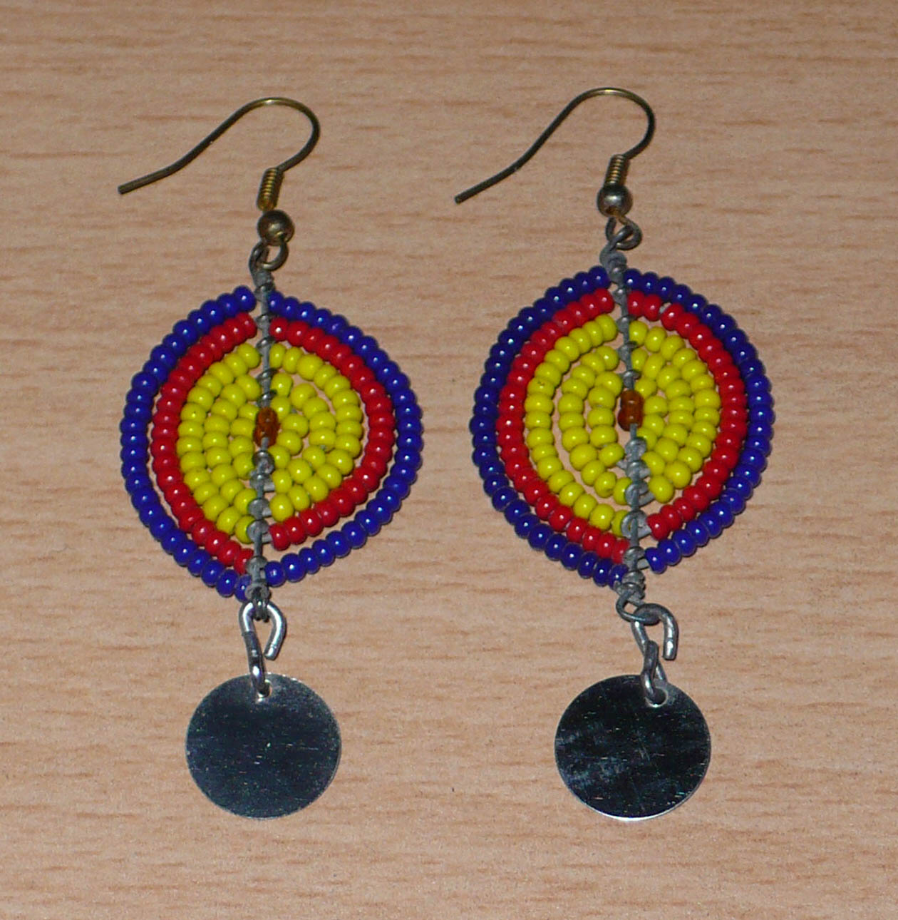 Boucles d'oreilles africaines en forme de boucliers Massai composées de perles de rocaille jaunes, rouges et bleu foncé ; elles se prolongent d'un disque métallique. Ces boucles d'oreilles mesurent 7 cm de long et 2,5 cm de large ; elles se portent sur des oreilles percées.