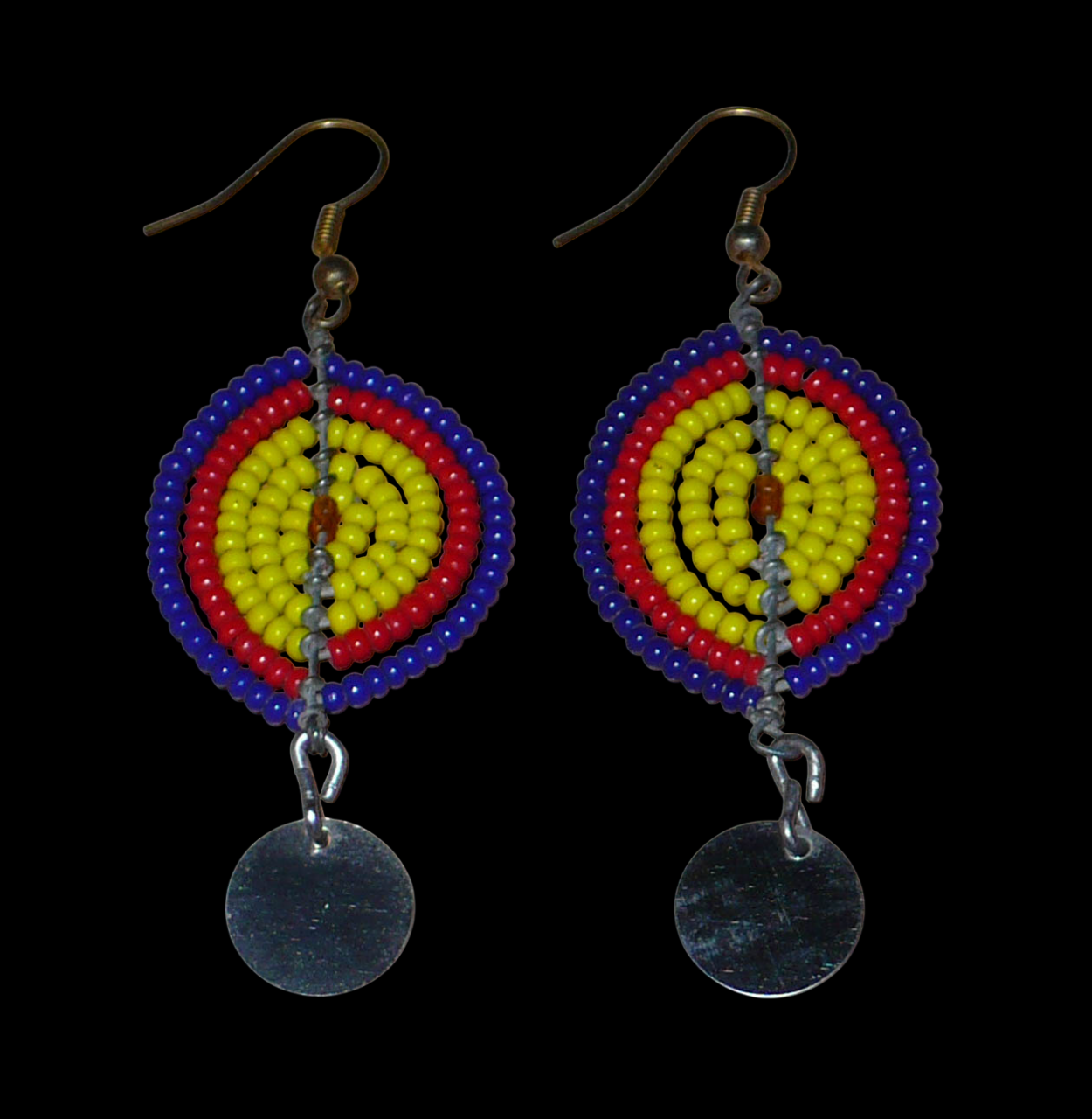 Boucles d'oreilles africaines en forme de boucliers Massai composées de perles de rocaille jaunes, rouges et bleu foncé ; elles se prolongent d'un disque métallique. Ces boucles d'oreilles mesurent 7 cm de long et 2,5 cm de large ; elles se portent sur des oreilles percées.