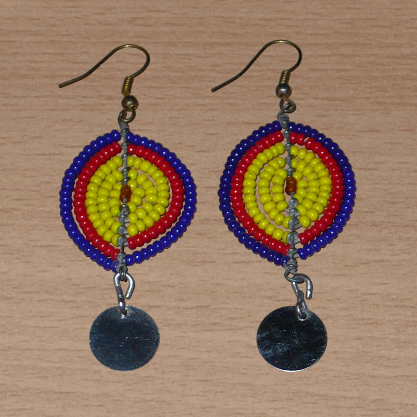 Boucles d'oreilles africaines en forme de boucliers Massai composées de perles de rocaille jaunes, rouges et bleu foncé ; elles se prolongent d'un disque métallique. Ces boucles d'oreilles mesurent 7 cm de long et 2,5 cm de large ; elles se portent sur des oreilles percées.