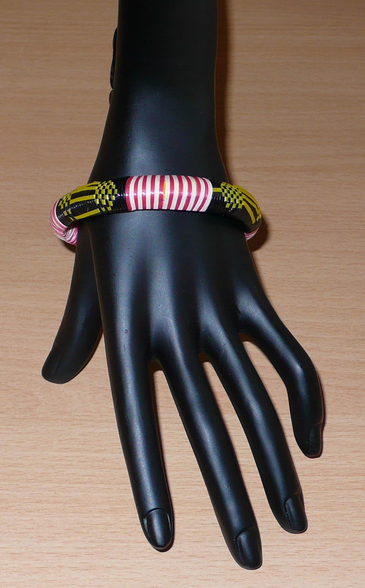 Bracelet africain fabriqué dans une optique eco-responsable avec des matériaux de récupération : il se compose de motifs ethniques tissés à partir de fines bandes de plastique recyclé noir, jaune, rouge et blanc.