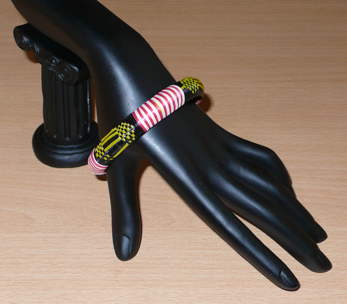 Bracelet africain fabriqué dans une optique eco-responsable avec des matériaux de récupération : il se compose de motifs ethniques tissés à partir de fines bandes de plastique recyclé noir, jaune, rouge et blanc.