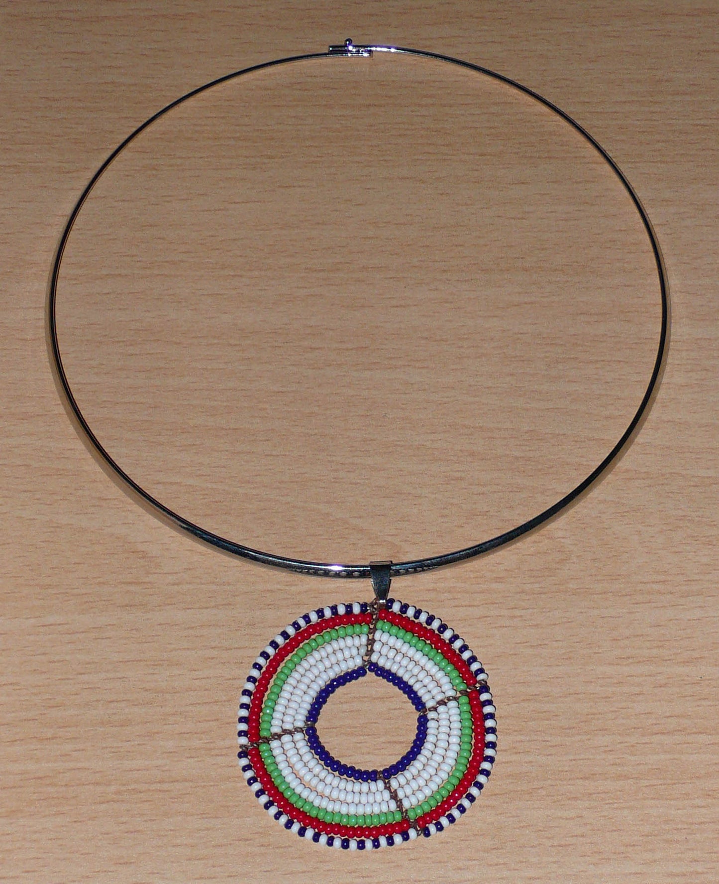 Collier ethnique contemporain composé d'un ras de cou en laiton orné d'un médaillon Massai fait de perles de rocaille blanches, vertes, rouges, bleu marine et noires.