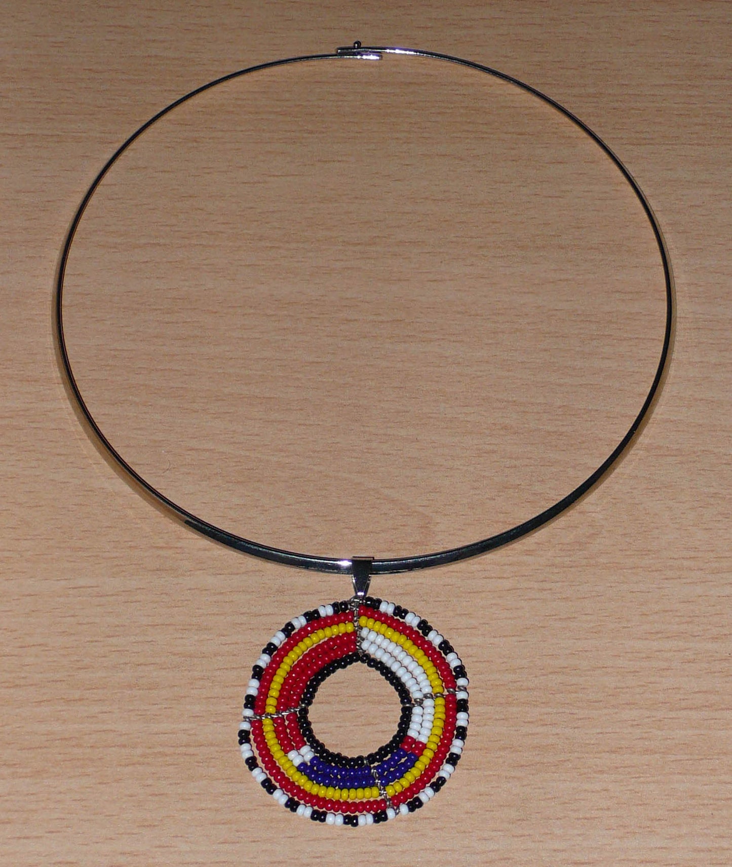 Collier ethnique contemporain se composant d'un ras de cou en laiton orné d'un pendentif rond à motifs Massai fait d'un tissage de perles de rocaille rouges, blanches, bleues, jaunes, noires et blanches.
