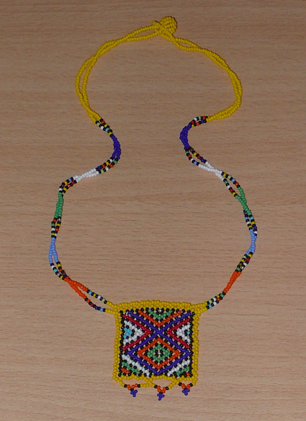 Collier africain Zoulou avec pendentif orné de motifs ethniques traditionnels. Il se compose de perles de rocaille jaunes et multicolores et s'attache avec un fermoir à boule.