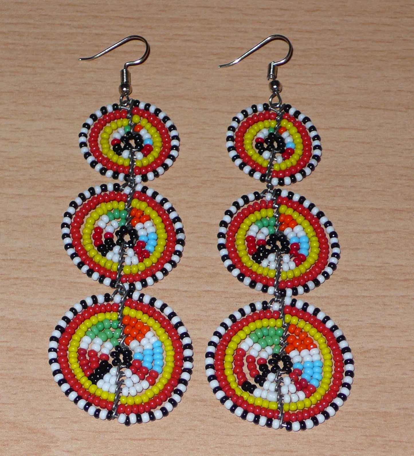 Longues boucles d'oreilles africaines de tradition Massai aux design et couleurs évoquant le style ethnique afro chic des années 70. Elles se composent de perles de rocaille multicolores glissées sur des fils de fer montés en forme de trois cercles superposés. Ces boucles d'oreilles mesurent 10.5 cm de long et 3 cm de large ; elles se portent avec des crochets en acier inoxydable sur des oreilles percées.