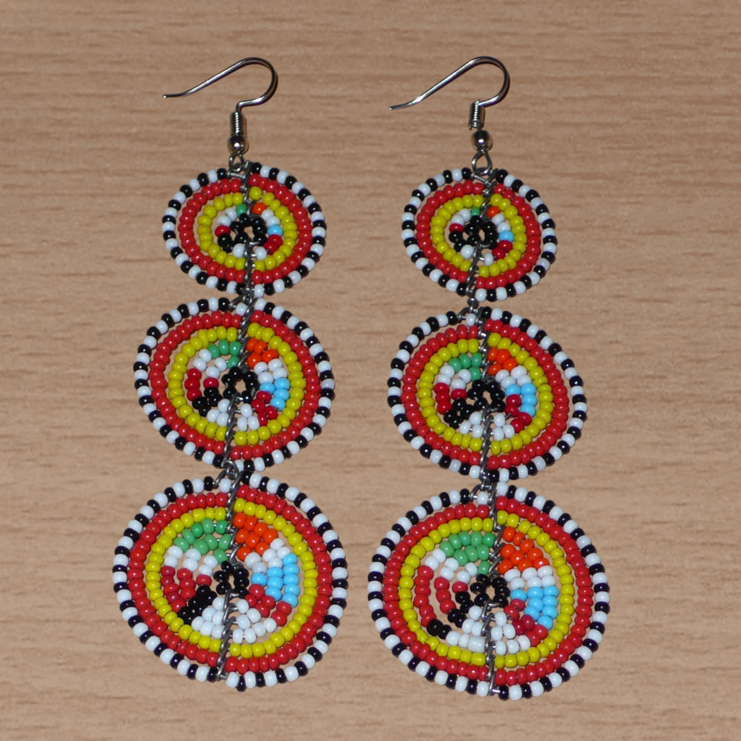Longues boucles d'oreilles africaines de tradition Massai aux design et couleurs évoquant le style ethnique afro chic des années 70. Elles se composent de perles de rocaille multicolores glissées sur des fils de fer montés en forme de trois cercles superposés. Ces boucles d'oreilles mesurent 10.5 cm de long et 3 cm de large ; elles se portent avec des crochets en acier inoxydable sur des oreilles percées.