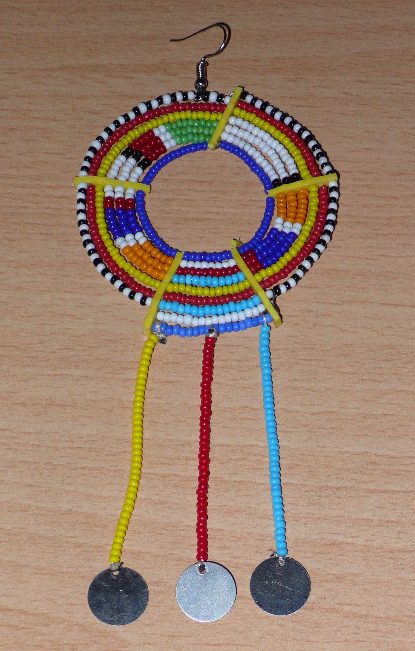 Mono boucle d'oreille africaine Massai à porter sur une seule oreille pour créer un effet d'asymétrie visuelle : elle se compose de perles de rocaille bleues et multicolores enfilées sur des fils de fer montés en cercles fixés sur des charnières en plastique recyclé jaune ; il se prolonge de franges perlées ornées de disques en métal. Surdimensionnée, cette mono boucle d'oreille à l'allure exotique mesure 15 cm de long.