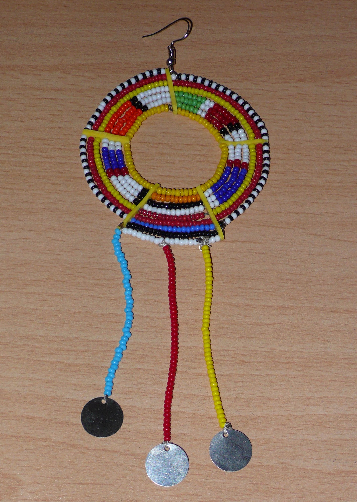 Mono boucle d'oreille à franges fabriquée au Kenya dans la tradition ethnique Massai. À porter sur une seule oreille pour un effet visuel asymétrique, ce bijou se compose de perles de rocaille multicolores glissées sur des fils de fer montés en cercles fixés sur des charnières en plastique recyclé jaune ; il se prolonge de franges perlées ornées de disques en métal. Surdimensionnée, elle mesure 16.5 cm de long et 6.5 cm de large, et se porte sur une oreille percée.
