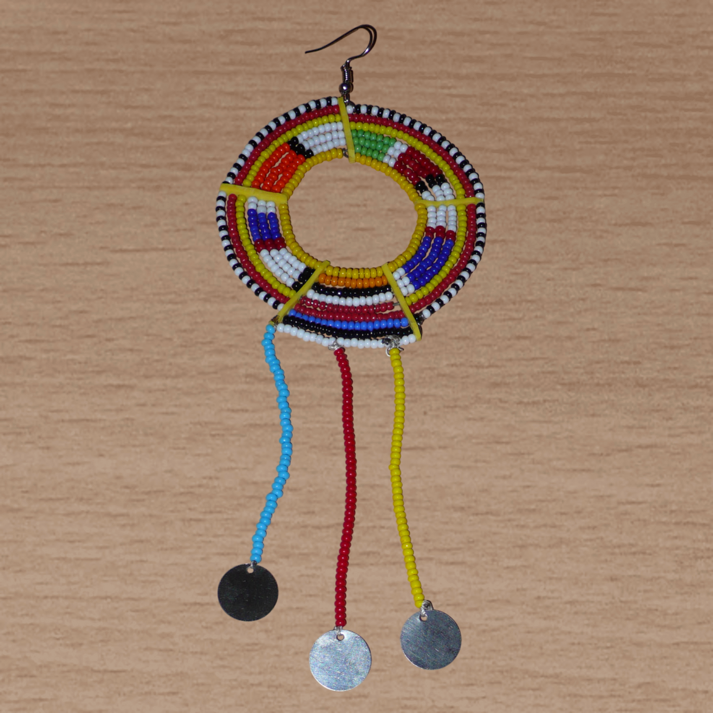 Mono boucle d'oreille à franges fabriquée au Kenya dans la tradition ethnique Massai. À porter sur une seule oreille pour un effet visuel asymétrique, ce bijou se compose de perles de rocaille multicolores glissées sur des fils de fer montés en cercles fixés sur des charnières en plastique recyclé jaune ; il se prolonge de franges perlées ornées de disques en métal. Surdimensionnée, elle mesure 16.5 cm de long et 6.5 cm de large, et se porte sur une oreille percée.