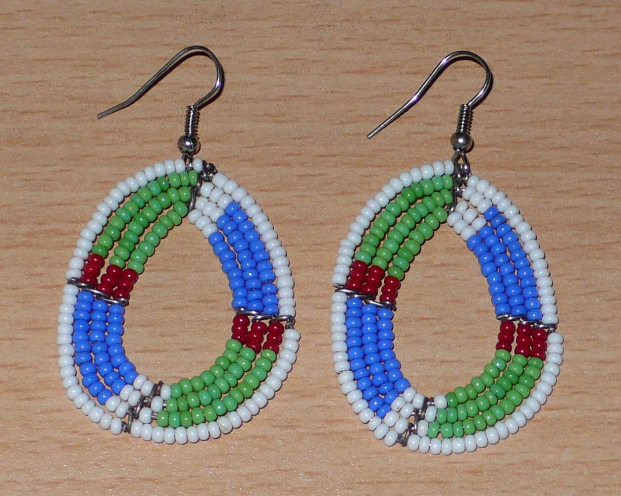 Boucles d'oreilles africaines Massai en forme de gouttes composées de perles de rocaille vertes, bleues, rouges et blanches. Elles mesurent 6 cm de long et 3,5 cm de large et se portent avec des crochets en acier inoxydable sur des oreilles percées.