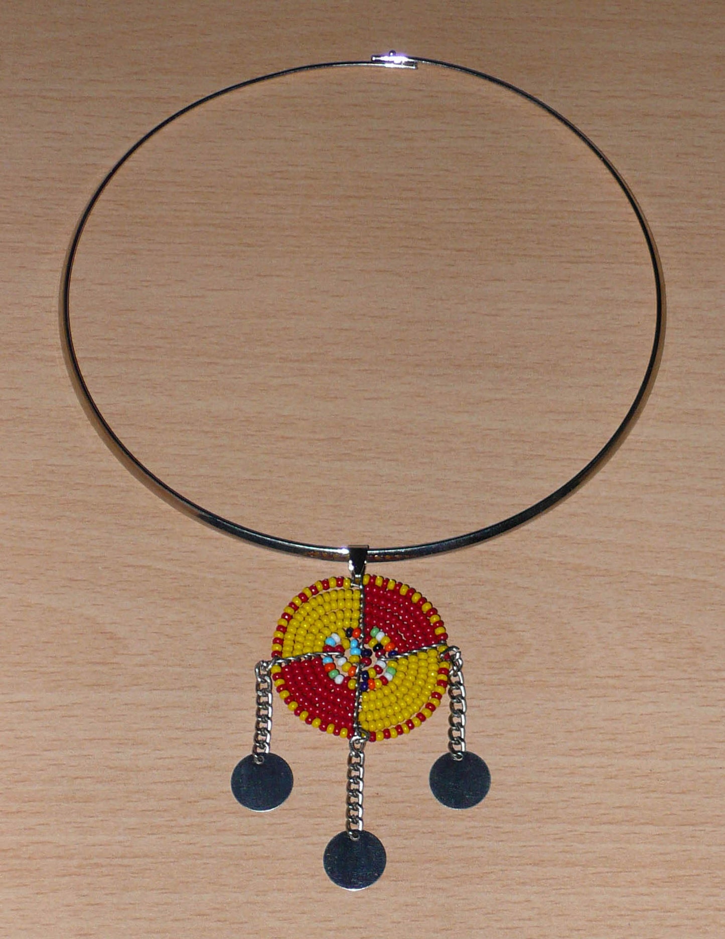 Bijou africain se composant d'un collier ras de cou en laiton rehaussé d'un pendentif Massai rond fait de perles de rocaille rouges, jaunes et multicolores, et prolongé de trois chaînettes ornées de disques métalliques.