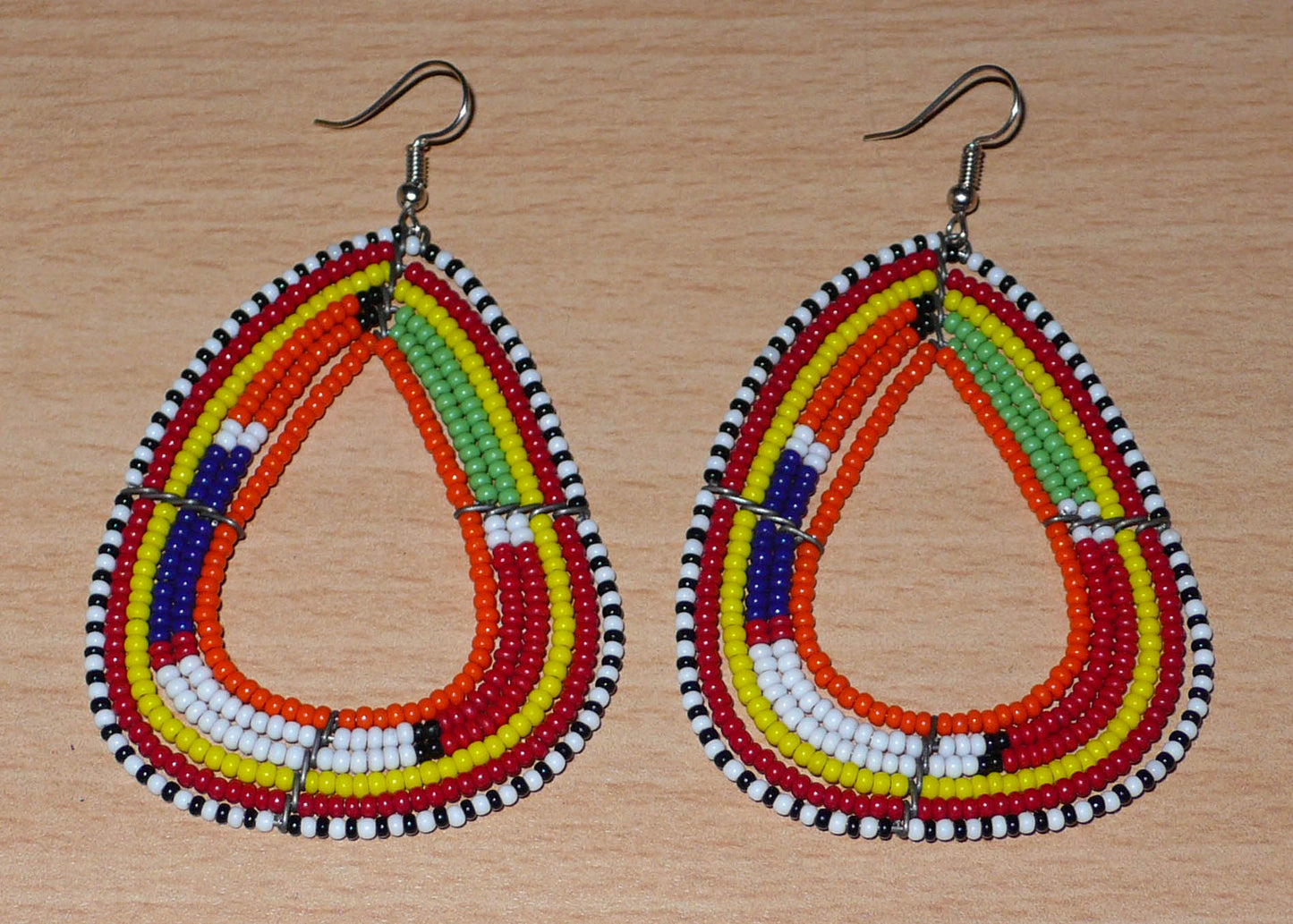 Boucles d'oreilles africaines traditionnelles à motifs ethniques Massai composées de perles de rocaille orange et multicolores enfilées sur six rangées de fils de fer montés en forme de triangles aux bords arrondis. Elles mesurent 8,5 cm de long et 5 cm de large et se portent avec des crochets en acier inoxydable sur des oreilles percées.