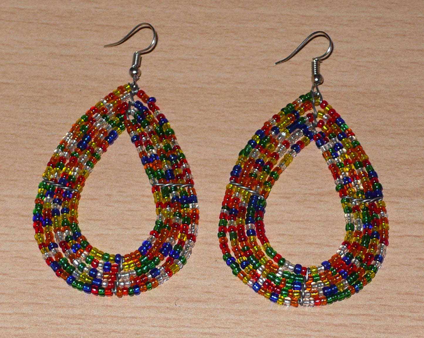 Boucles d'oreilles ethniques composées d'un mélange de perles de rocaille translucides multicolores enfilées sur cinq rangées de fils de fer montés en forme de gouttes. Elles mesurent 8 cm de long et 4,5 cm de large, et se portent avec des crochets sur des oreilles percées.