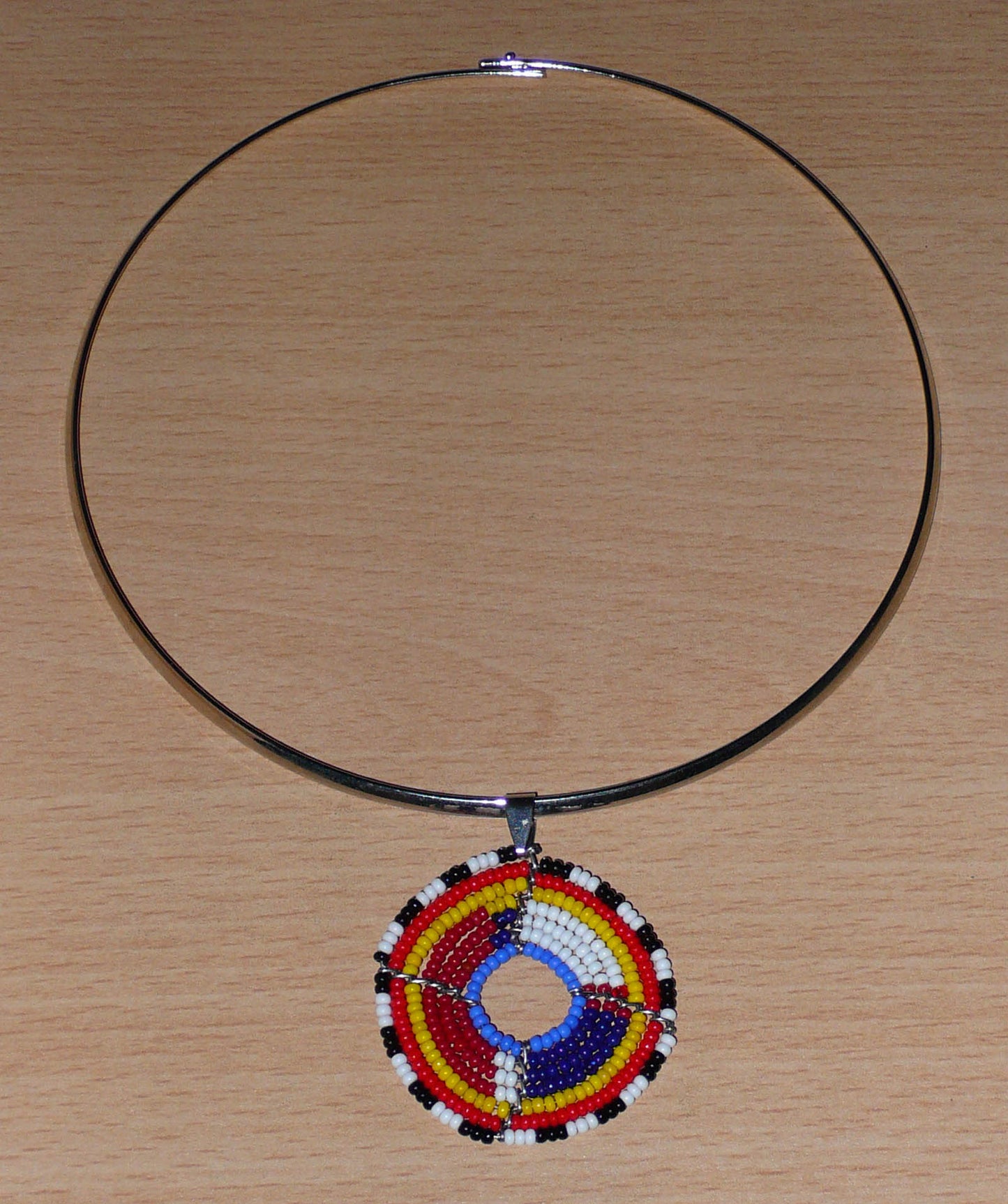 Collier africain composé d'un ras de cou en laiton orné d'un petit médaillon fait d'un tissage de perles de rocaille. Il mesure 18,5 cm de long, la circonférence du ras de cou est de 42,5 cm et son diamètre de 13,5 cm ; le médaillon mesure 4,5 cm de long et de large.