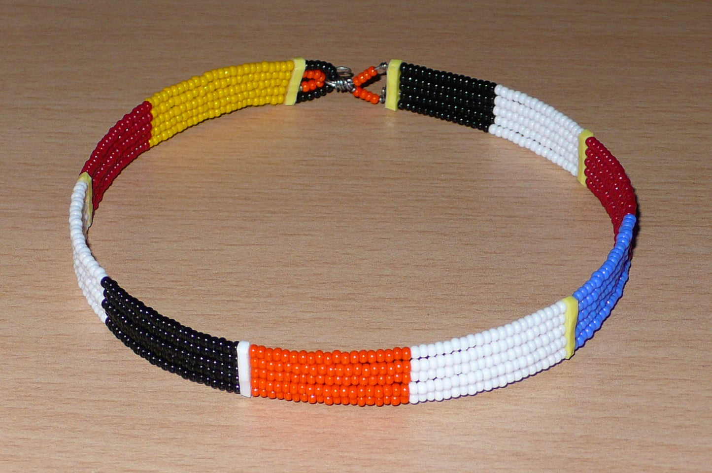 Collier ras de cou africain Massai fait de cinq rangées de perles de rocaille orange, noires, blanches, rouges et jaunes. Sa longueur autour du cou est de 35,5 cm et sa largeur de 1 cm ; il s'attache avec un crochet.