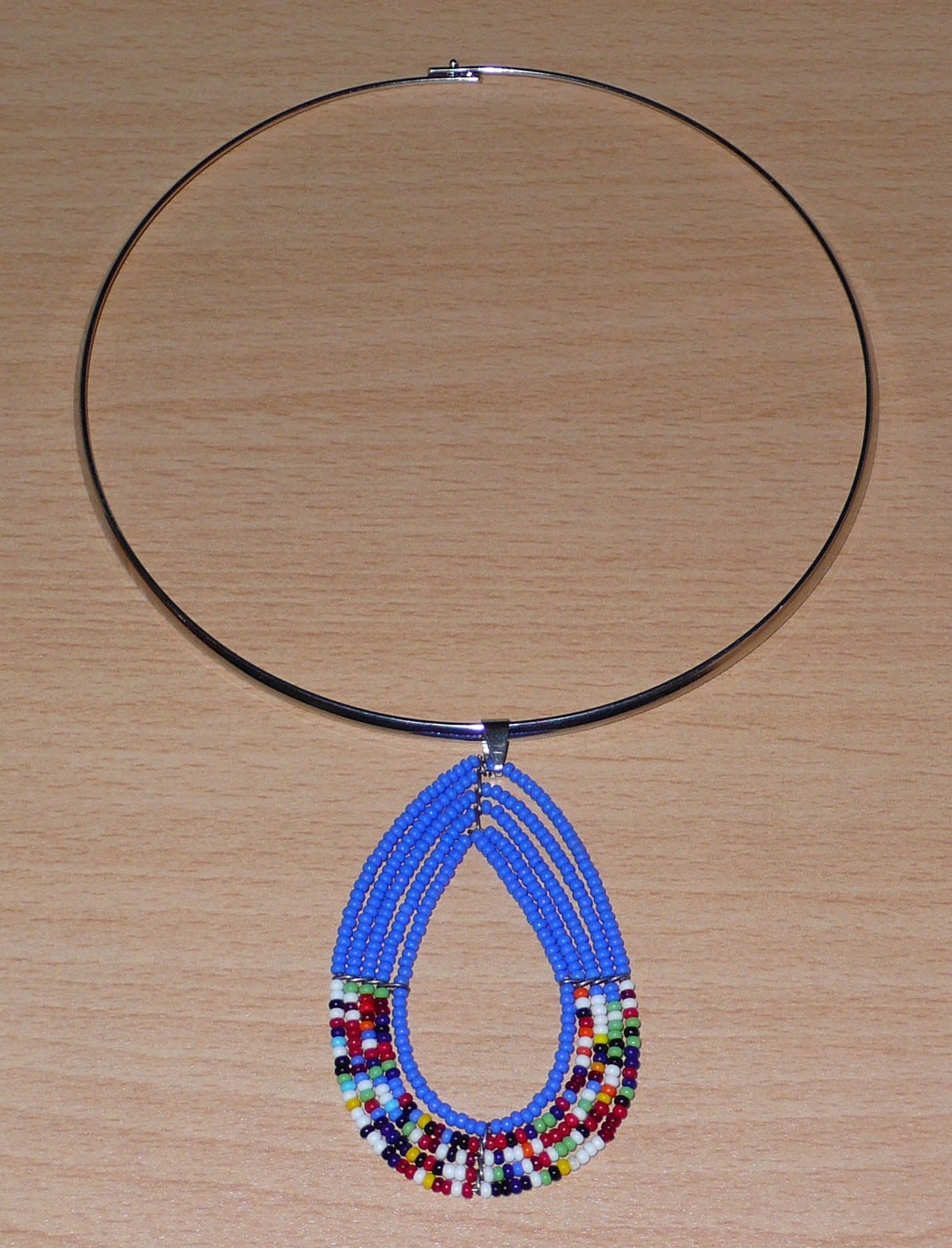 Collier ras de cou bleu et multicolore en laiton orné d'un long pendentif fait de perles de rocaille enfilées sur cinq rangées de fils de fer montés en forme de goutte. Il mesure 21 cm de long, la circonférence du ras de cou est de 4,5 cm avec un diamètre de 13,5 cm ; le pendentif mesure 7 cm de long et 4,5 cm de large.
