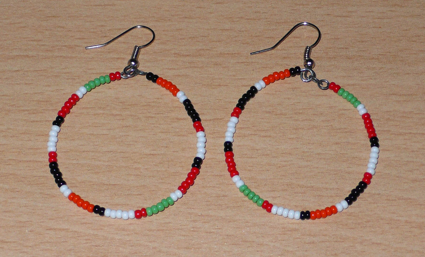 Créoles aux couleurs ethniques Massai faites de perles de rocaille rouges, vertes, noires, blanches et orange glissées sur un brin de fils de fer monté en cercle. Elles mesurent 6 cm de long et 4 cm de large, et se portent avec des crochets en acier inoxydable sur des oreilles percées.