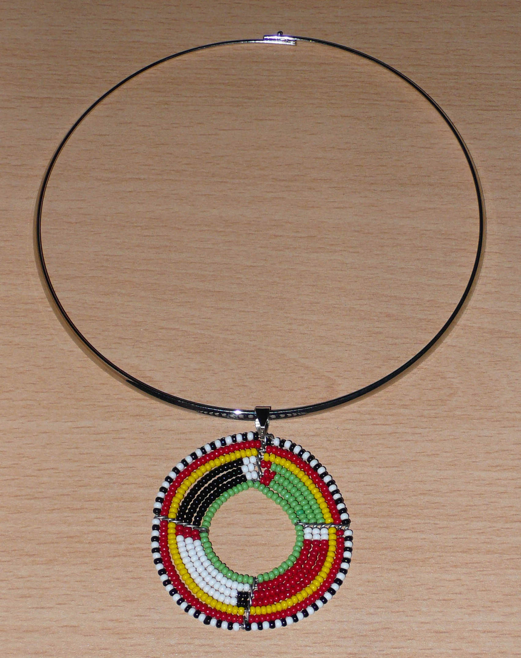 Bijou africain composé d'un collier ras de cou en laiton orné d'un médaillon Massai fait de perles de rocaille rouges, vertes, jaunes, blanches et noires enfilées sur sept rangées de fils de fer montés en cercles. Il mesure 19 cm de long, la circonférence du ras de cou en laiton est de 42,5 cm et 13,5 cm de diamètre ; le pendentif mesure 5,5 cm de long et de large.