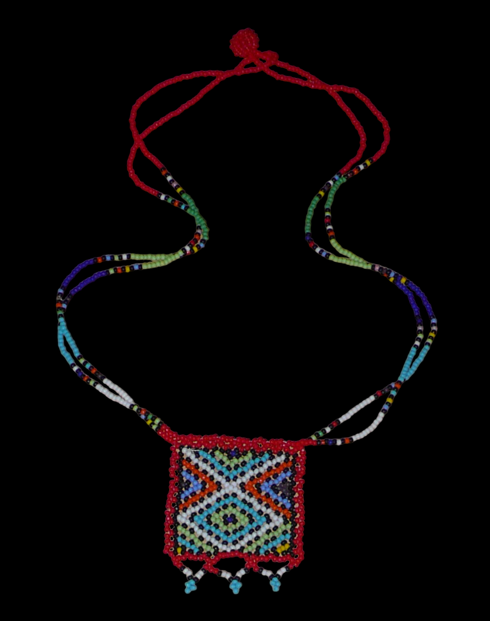 Collier africain composé de perles de rocaille rouges agrémenté d'un pendentif carré aux motifs multicolores zoulous. Il mesure 27,5 cm en partant de la nuque jusqu'à la base du pendentif. Le pendentif mesure 5,5 cm sur 4,5 cm. Le collier s'attache avec un fermoir à boule. Timeless Fineries
