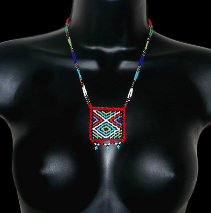 Collier africain composé de perles de rocaille rouges agrémenté d'un pendentif carré aux motifs multicolores zoulous. Il mesure 27,5 cm en partant de la nuque jusqu'à la base du pendentif. Le pendentif mesure 5,5 cm sur 4,5 cm. Le collier s'attache avec un fermoir à boule. Photographié sur un mannequin. Timeless Fineries